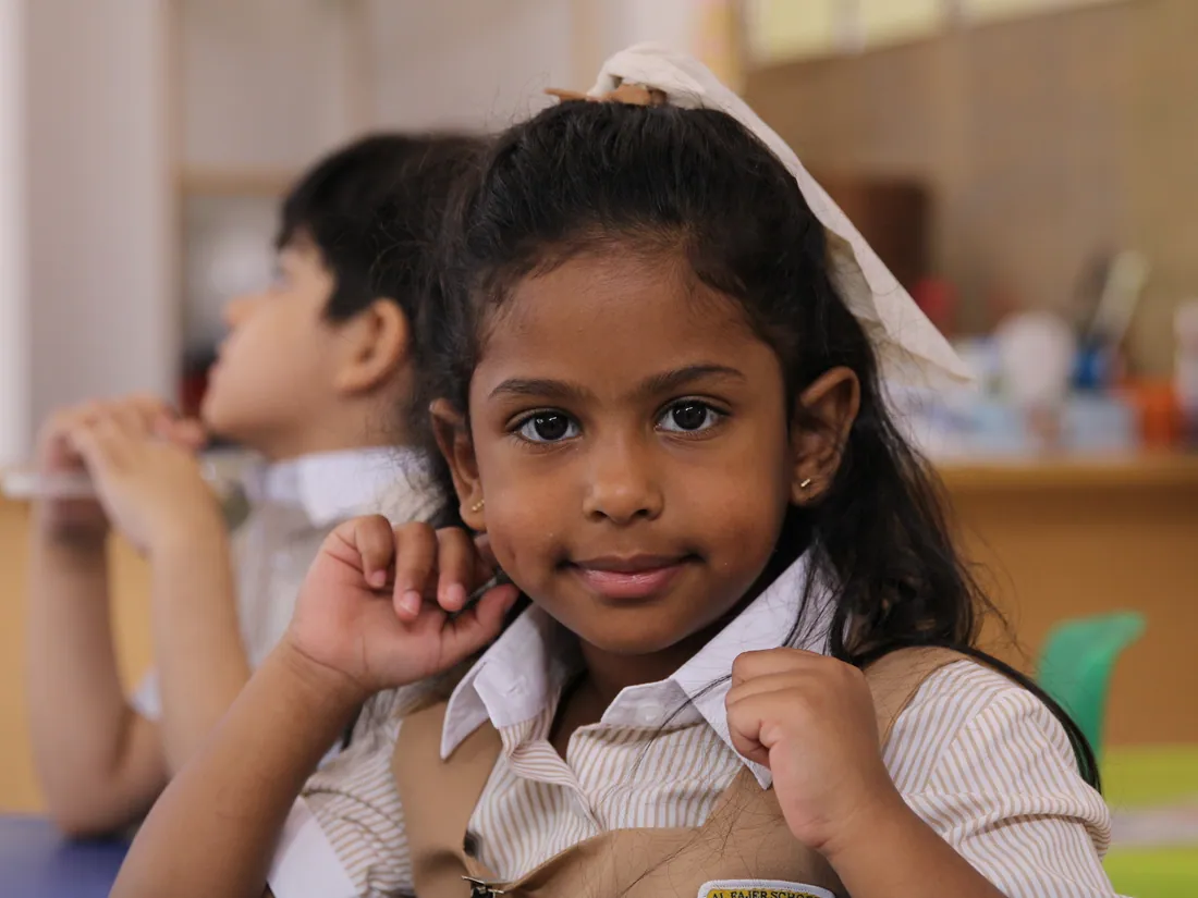 A smiling AFS pupil with a white hair bow at her desk, classmates behind her