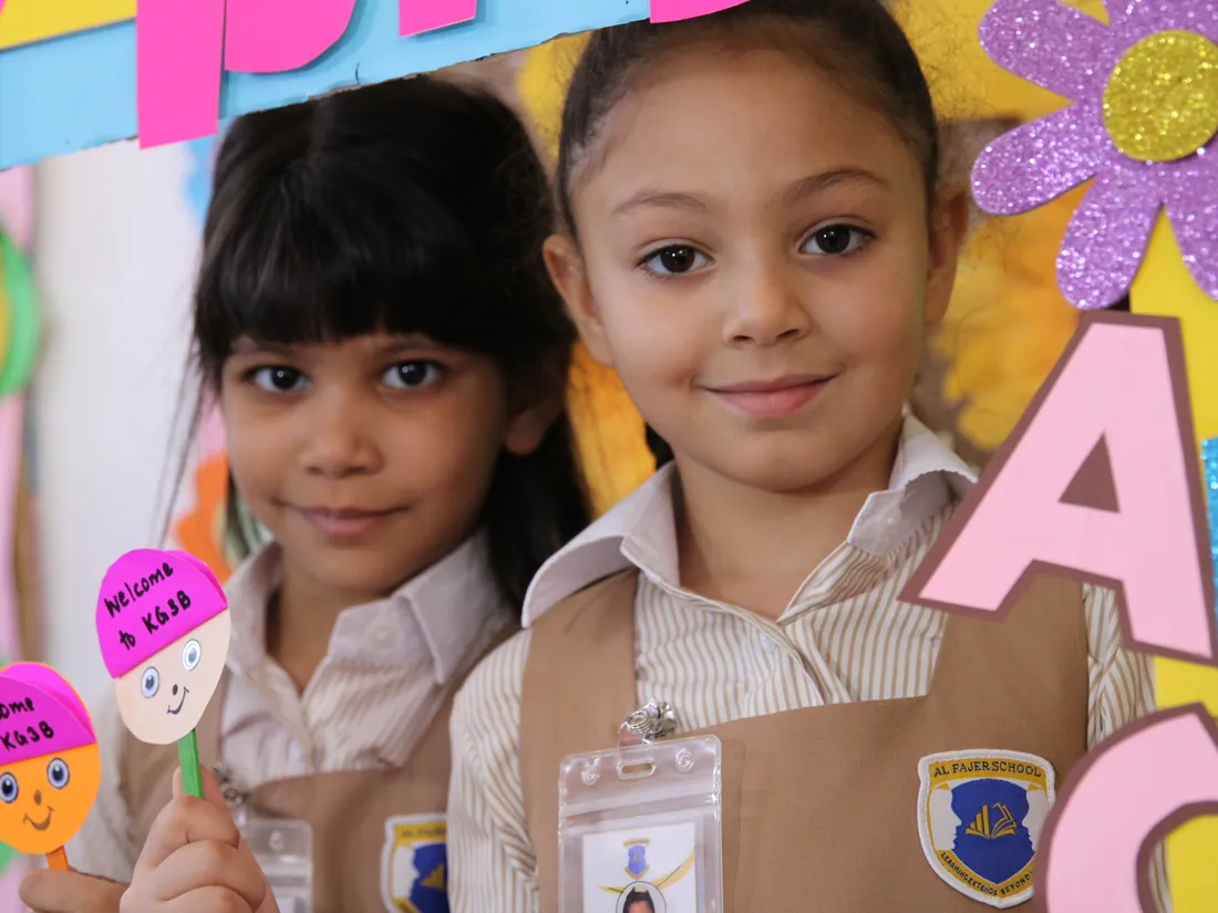 Two smiling AFS pupils in uniform, the Al Fajer crest on their pinafores, holding welcome crafts
