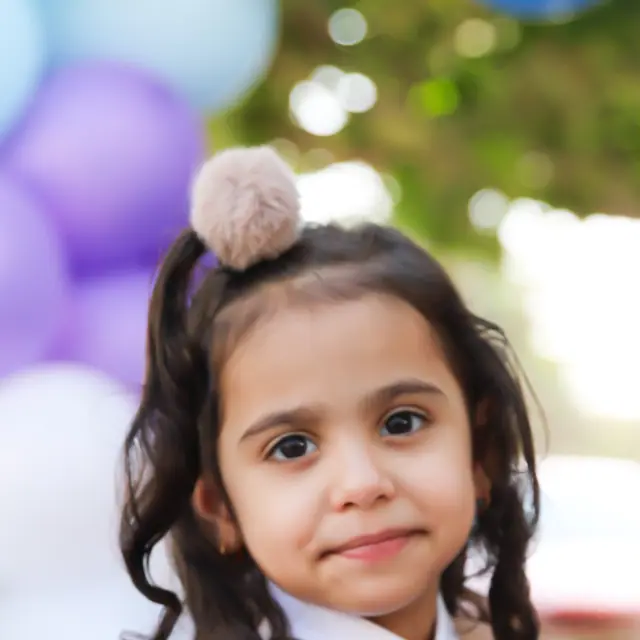 A young girl with pom-pom pigtails smiling softly in front of a pastel balloon arch