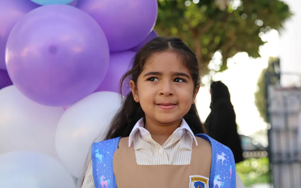 A young pupil with a unicorn backpack smiling shyly beside the welcome balloons